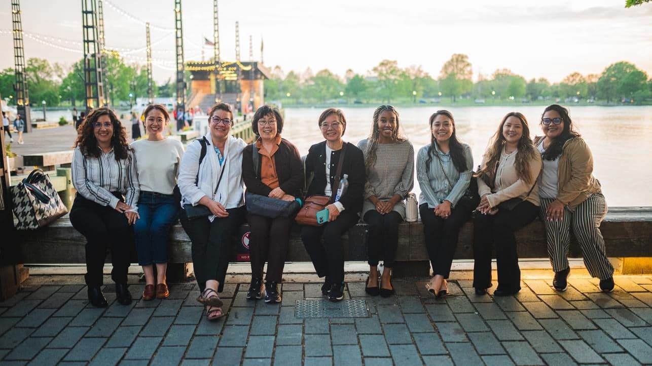 A group of nine people smiling as they sit together on a stone wall overlooking a lake at sunset.