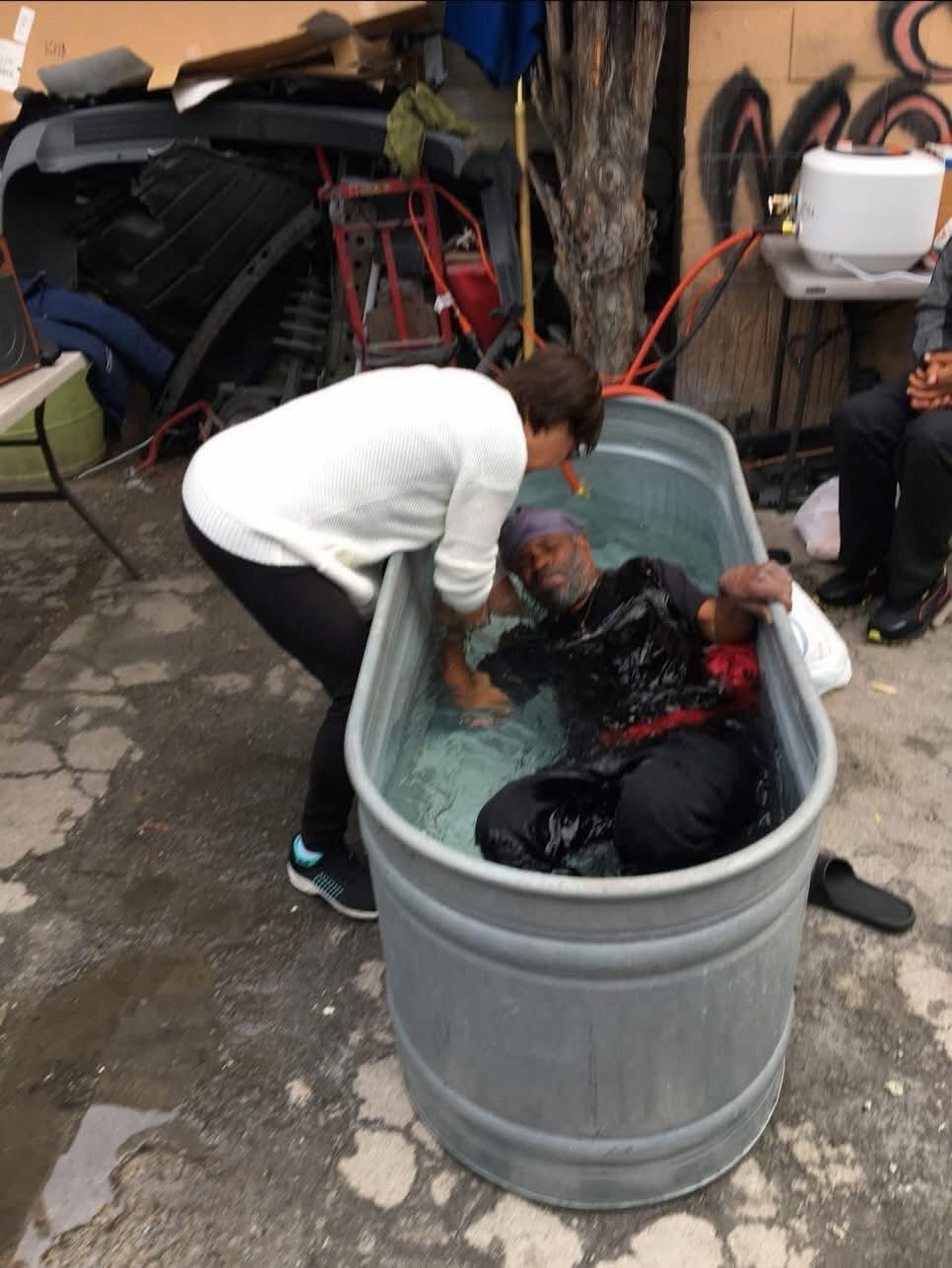 A person in a white sweater assists another individual sitting in a large metal stock tank filled with water outdoors.