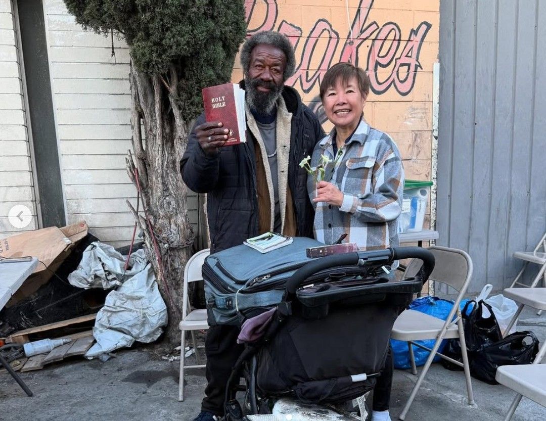 A smiling person and an individual holding a Bible stand together outside in front of a building with graffiti.