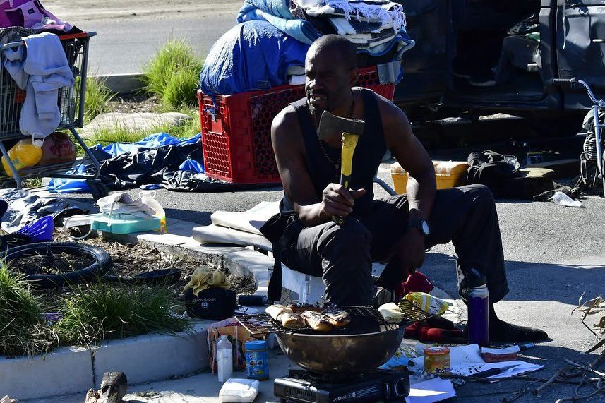 A person sits in an outdoor area surrounded by belongings, holding a small axe while cooking food on a portable grill.