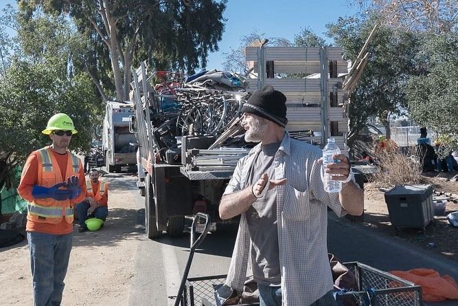 A worker in a safety vest stands near a truck filled with scrap metal while another person holds a water bottle nearby.