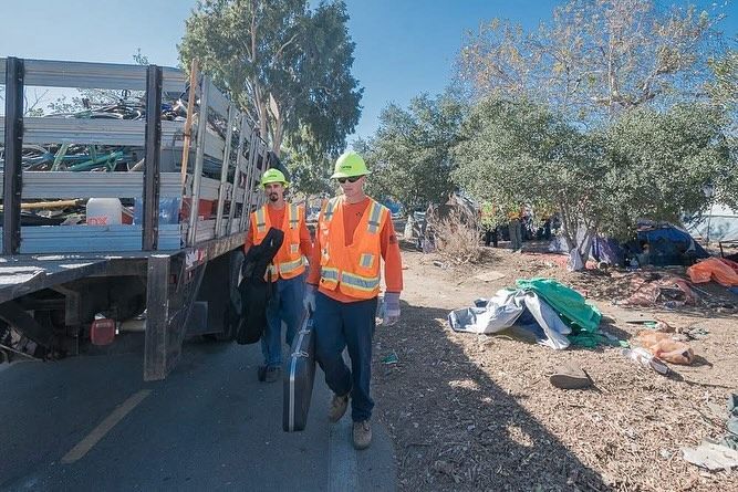 Two workers in high-visibility orange vests and hard hats walk past a truck loaded with debris near a roadside encampment.
