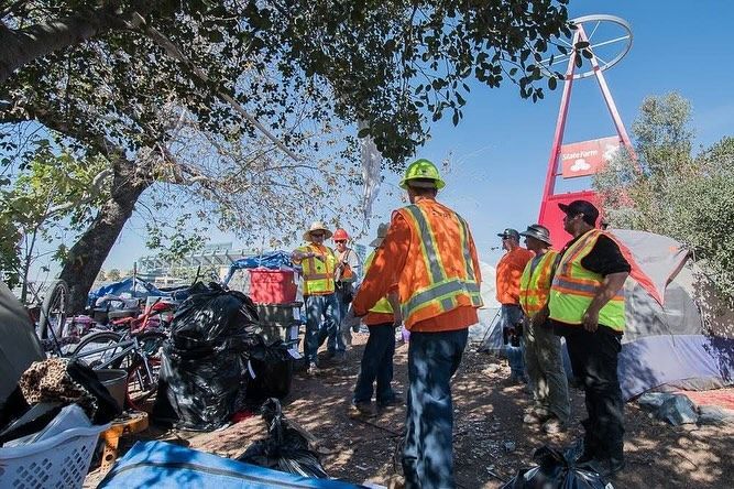 Workers in neon safety vests stand near a pile of trash bags and a red structure under trees during an outdoor cleanup.