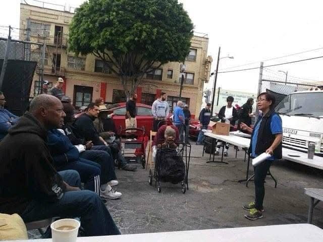 A woman stands and speaks to a seated group gathered outdoors in an urban area with buildings and a white vehicle nearby.