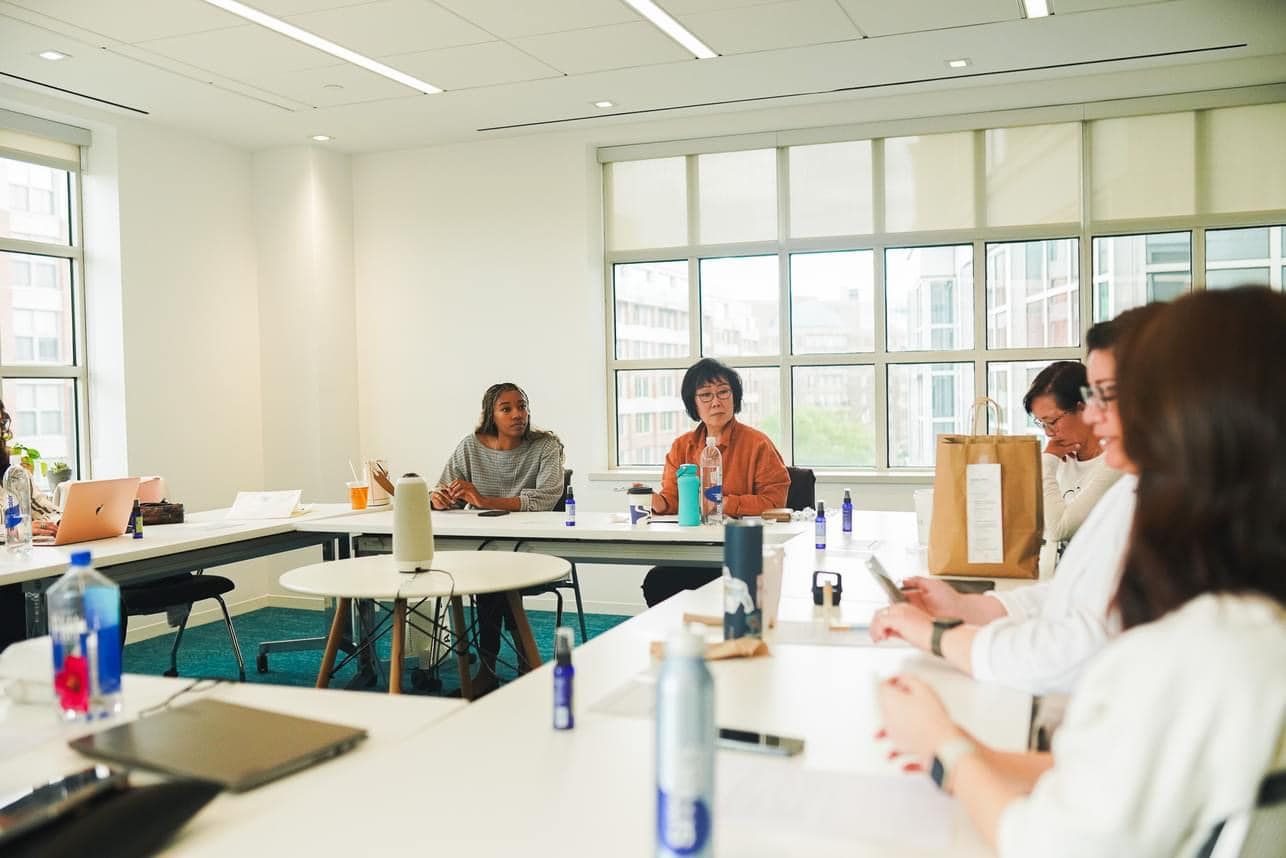 A group of people sit around a brightly lit conference table during a meeting in a modern, window-filled office.