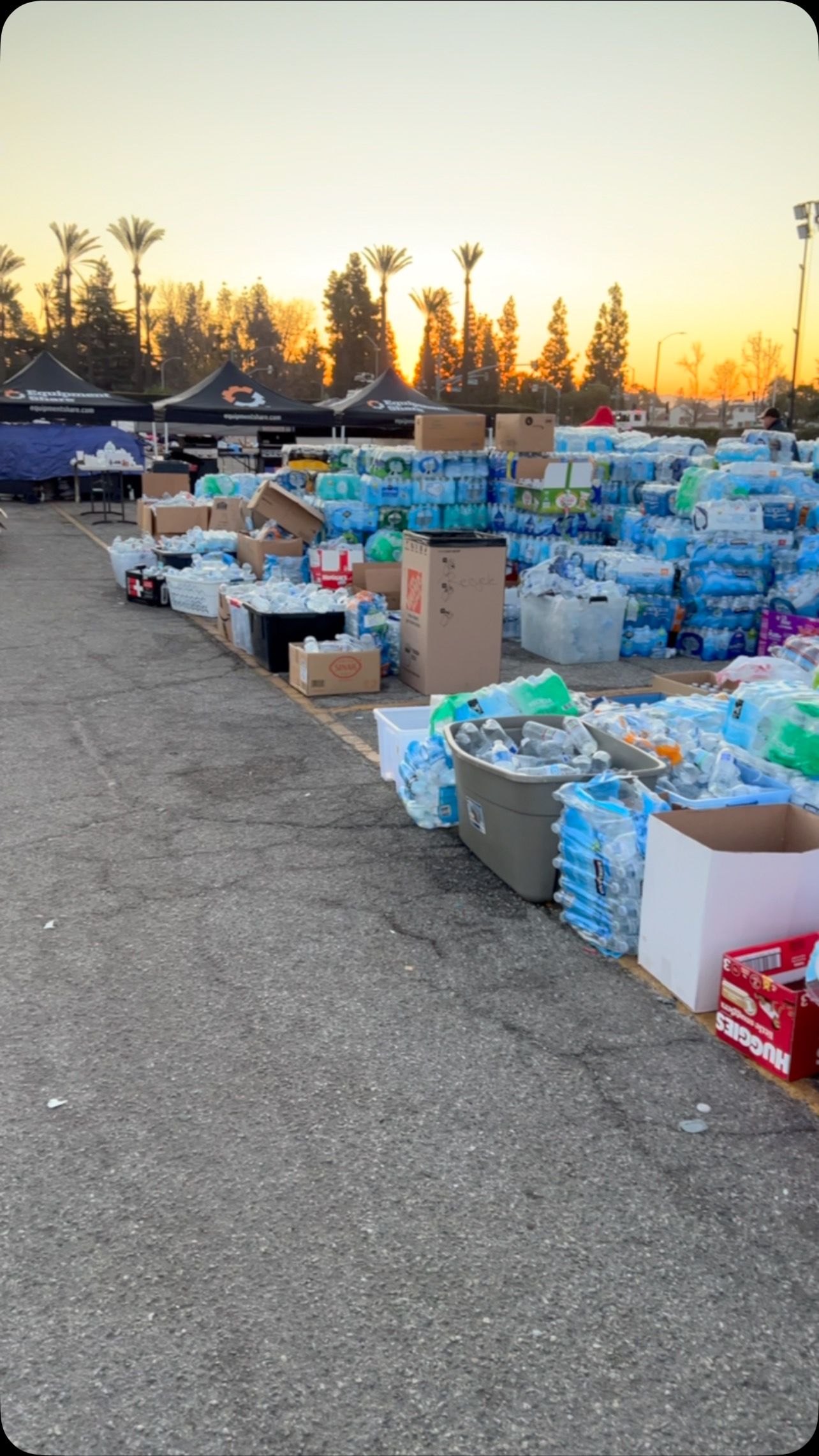 Large piles of water bottles in crates and boxes on an asphalt lot at sunset, likely representing a donation drive.