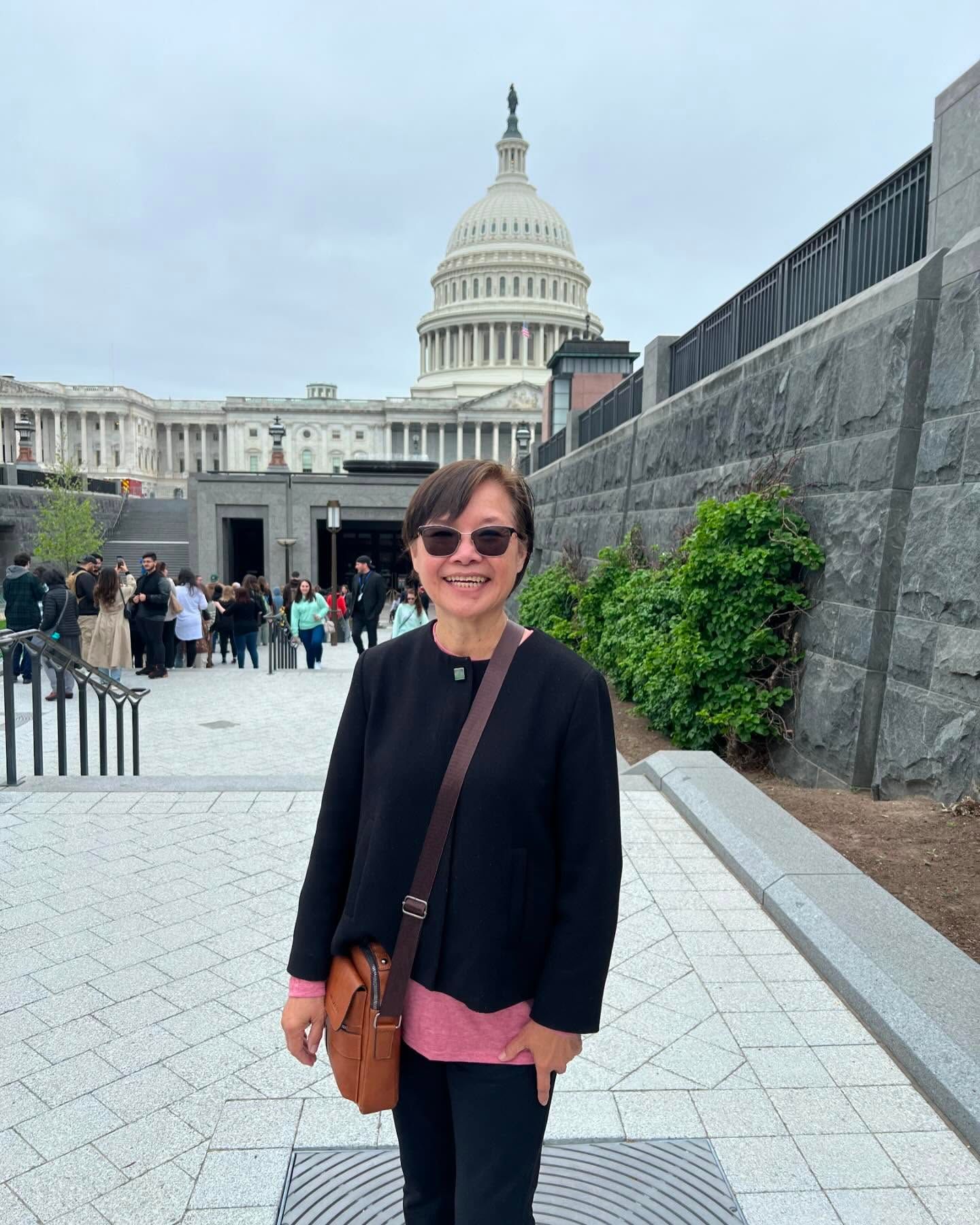 A person in a black jacket and pink shirt stands outside the US Capitol building, smiling while wearing sunglasses.