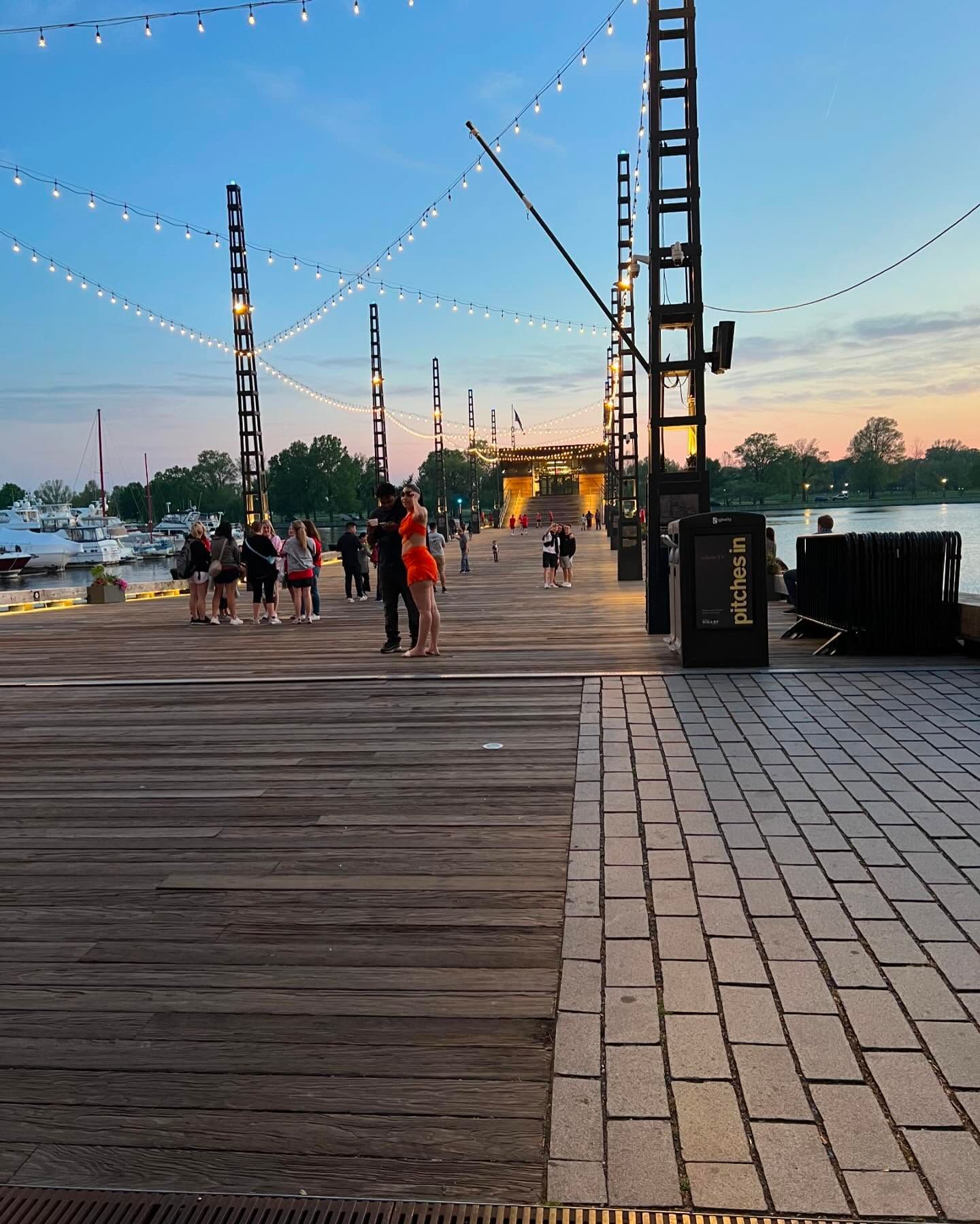People walk along a wood and brick pier at sunset, lit by overhead string lights, with boats docked in the background.