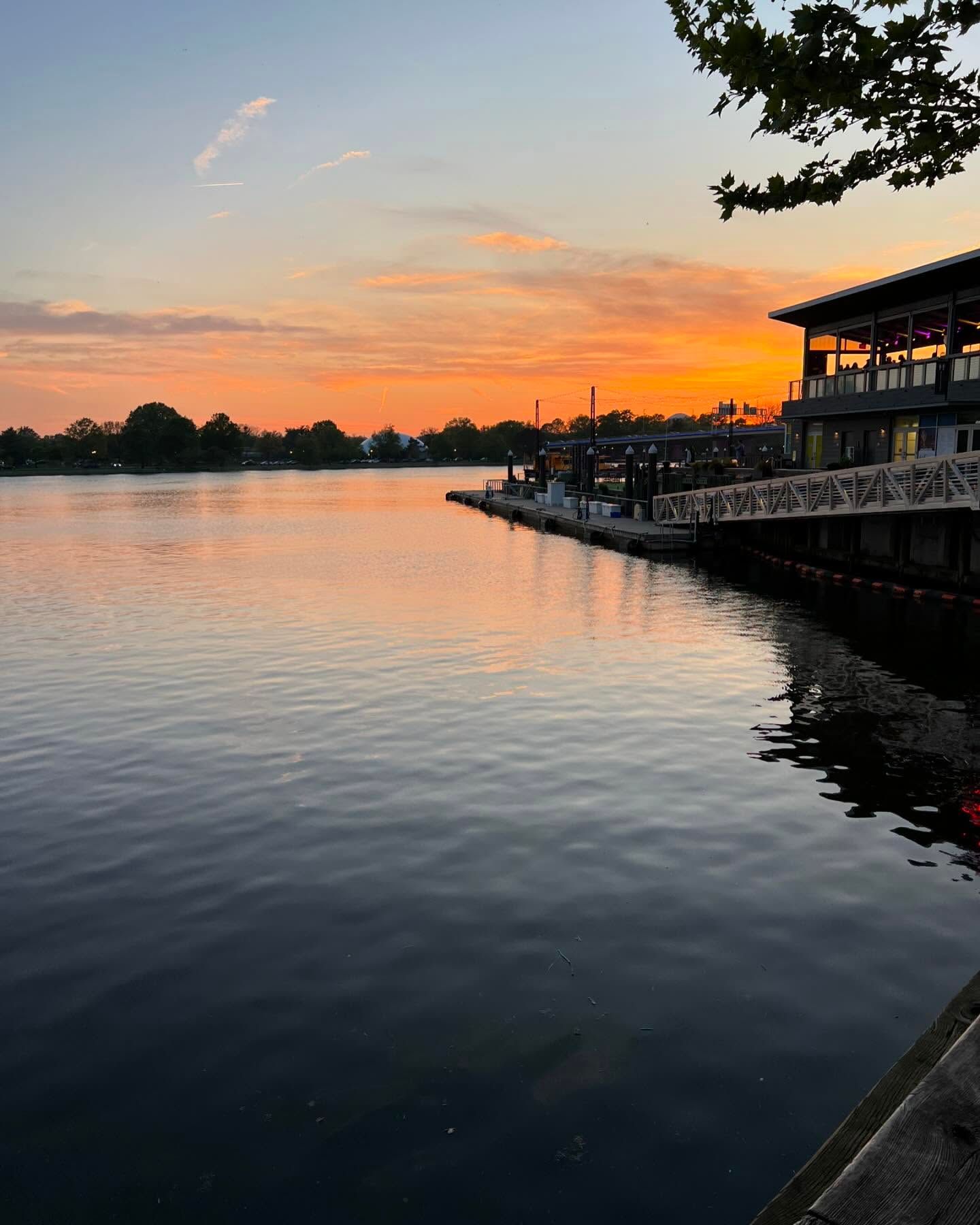 A vibrant orange and purple sunset over a calm river, featuring a wooden pier with a building on the right side.