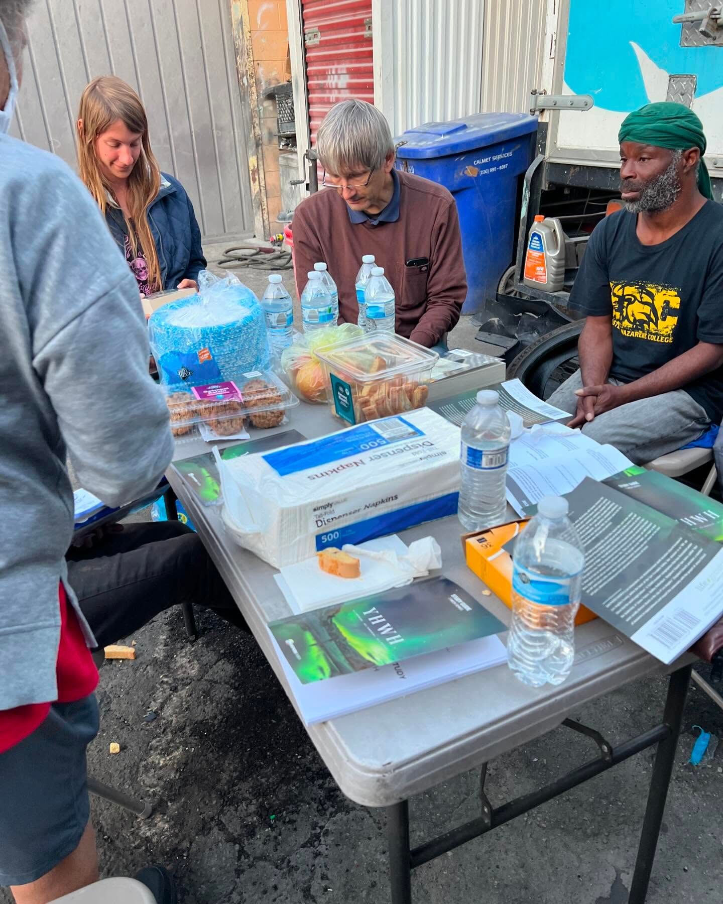 Four people sit around a folding table with snacks and water bottles in an outdoor setting.