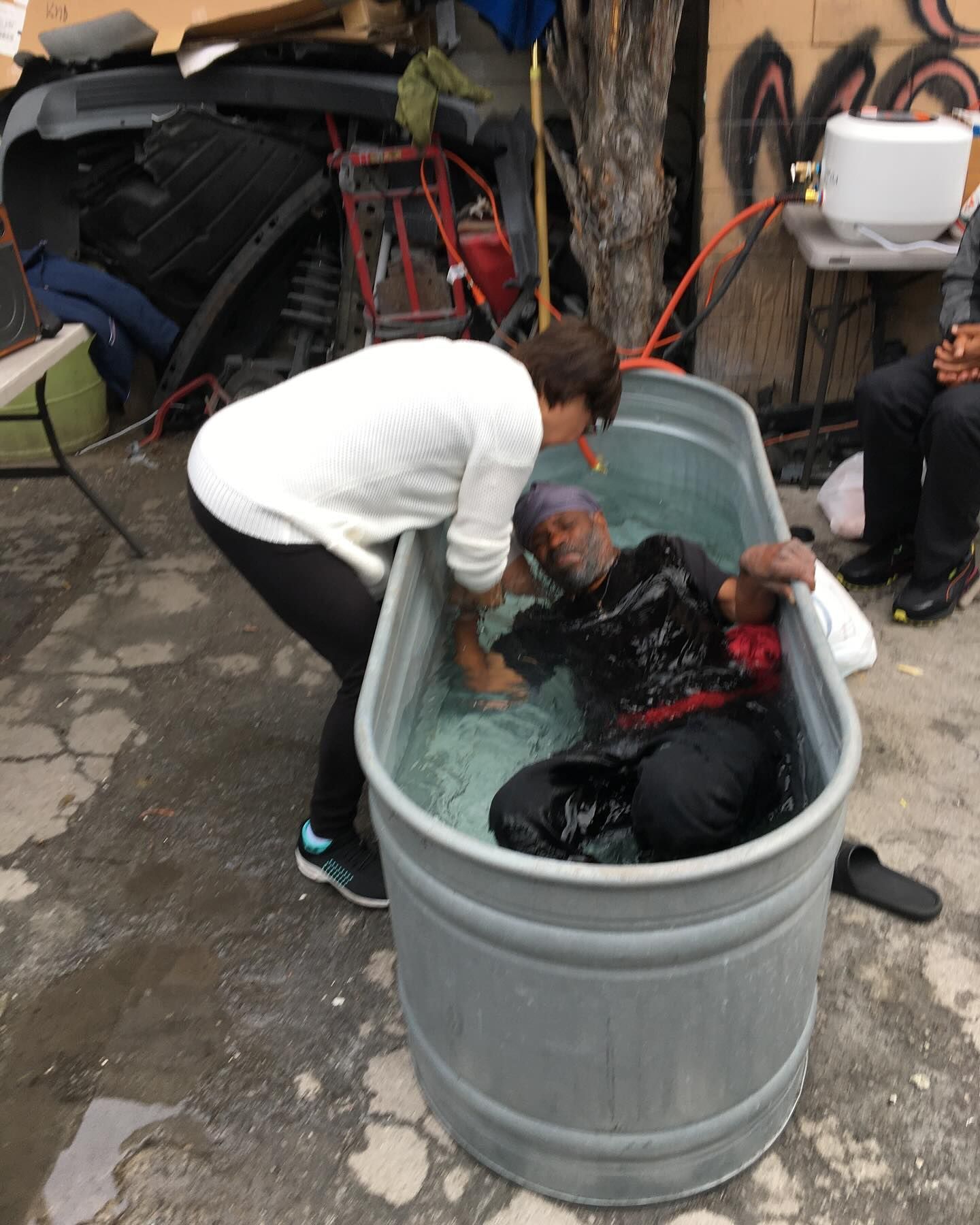 A person in a white sweater assists another individual, submerged in a metal trough filled with water, outdoors.