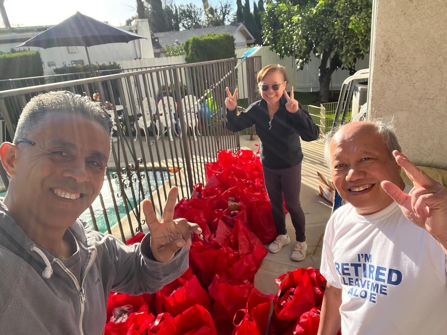Three people smile and make peace signs while standing behind a large row of red bags on a sunny outdoor patio.