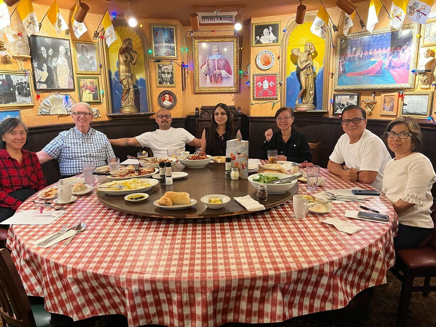 Eight people sit around a round, red-and-white checkered table in a restaurant decorated with framed art and statues.