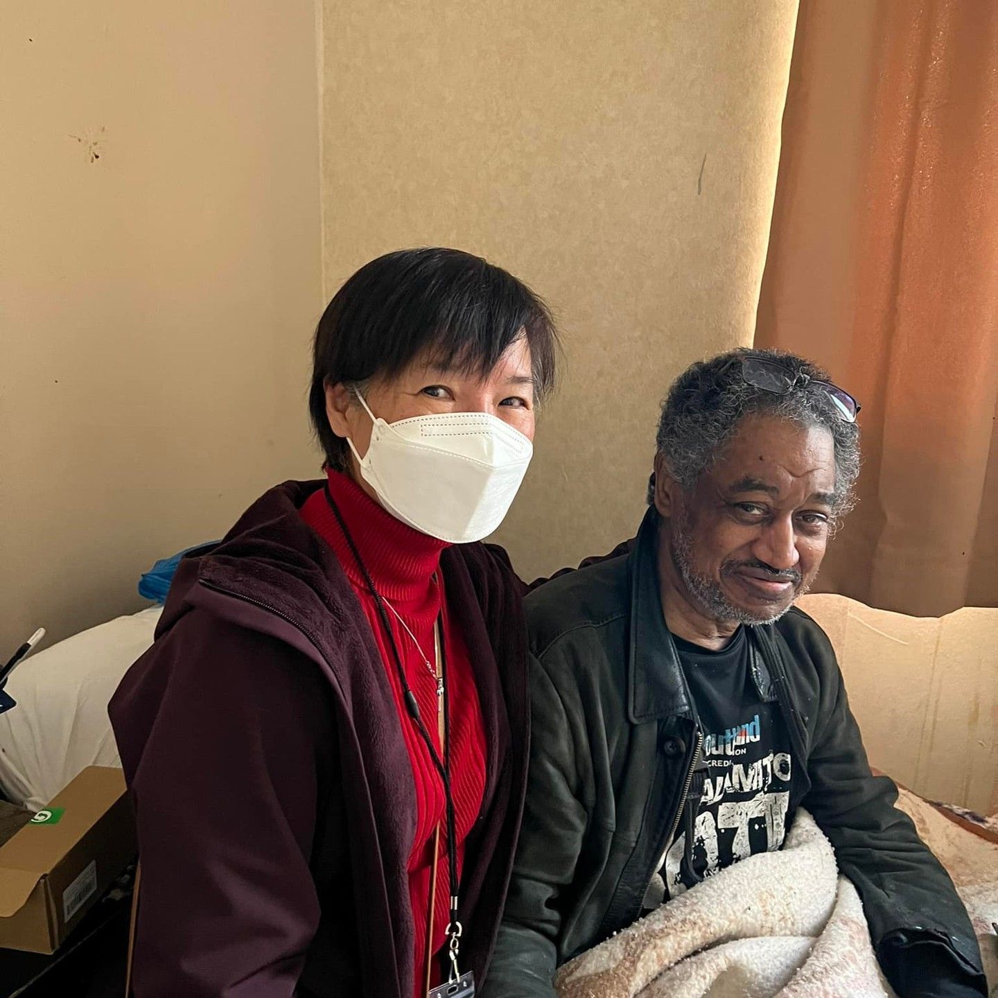 Two people smile together in an indoor setting; one wears a white mask and red sweater, the other a black jacket and blanket.