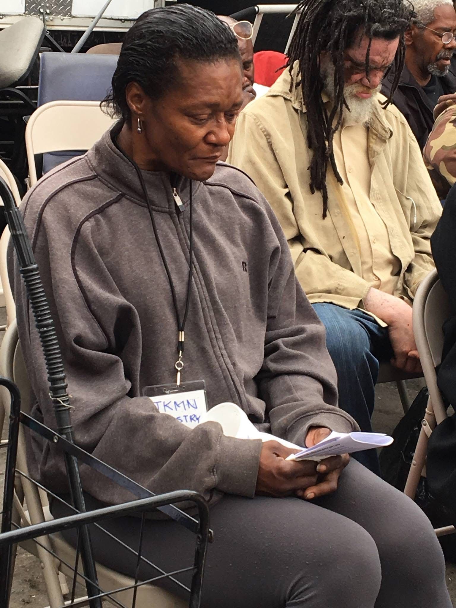 People sit in chairs, looking down at papers in a quiet, indoor setting.