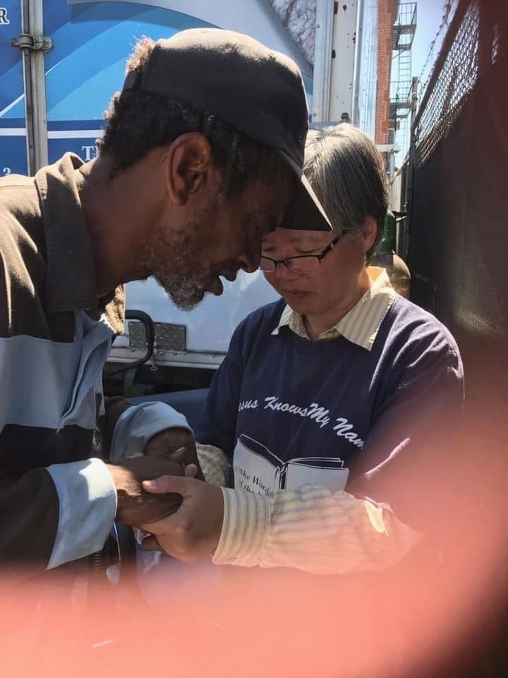 A person in a baseball cap gently holds the hands of another person, both with heads bowed in a moment of connection.