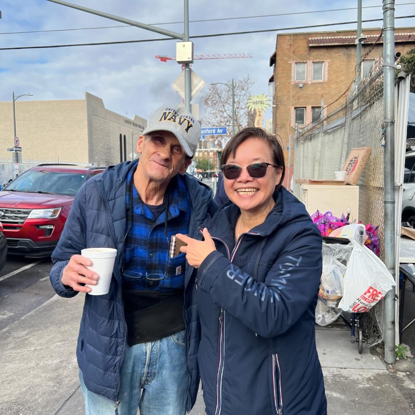 A person in a Navy hat and blue jacket smiles while holding a cup next to another person pointing toward them.