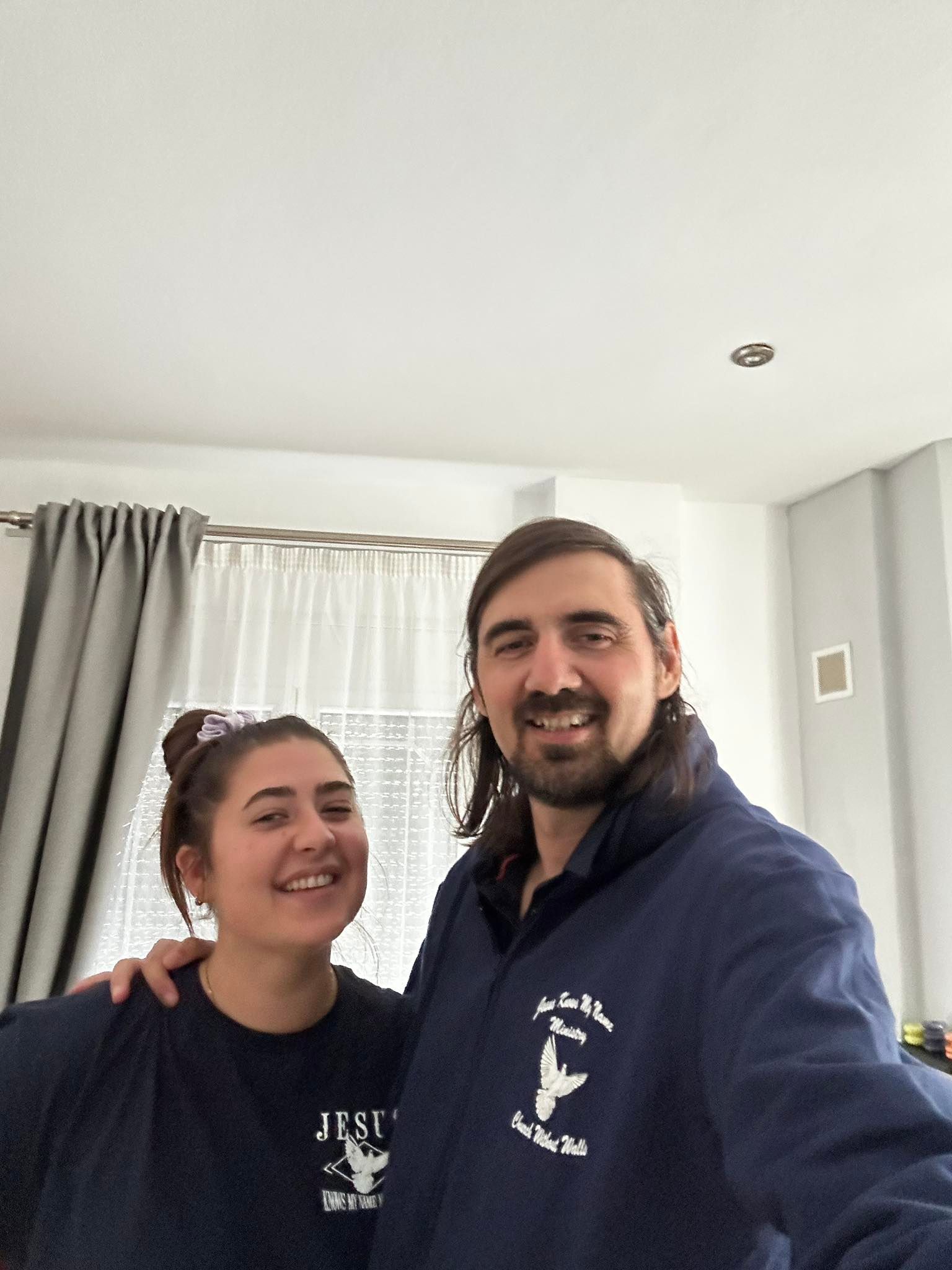 Two smiling people in matching navy blue clothing pose for a selfie in front of a window with light grey curtains.
