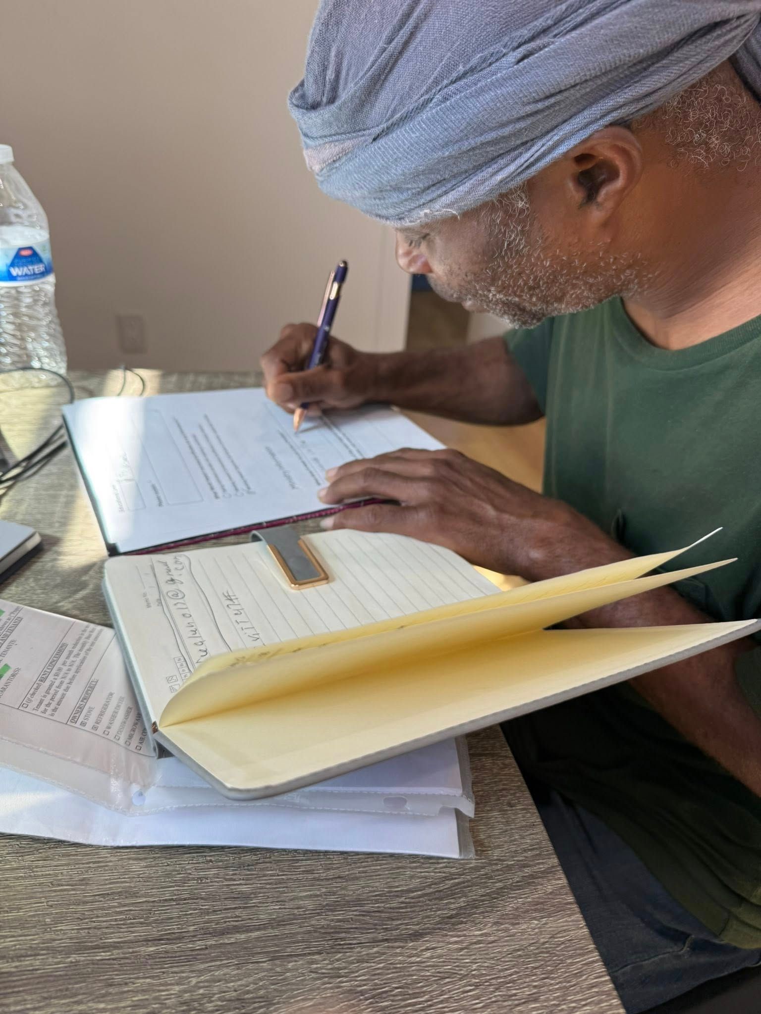A person wearing a head covering sits at a table, writing in an open notebook with a pen next to a water bottle.