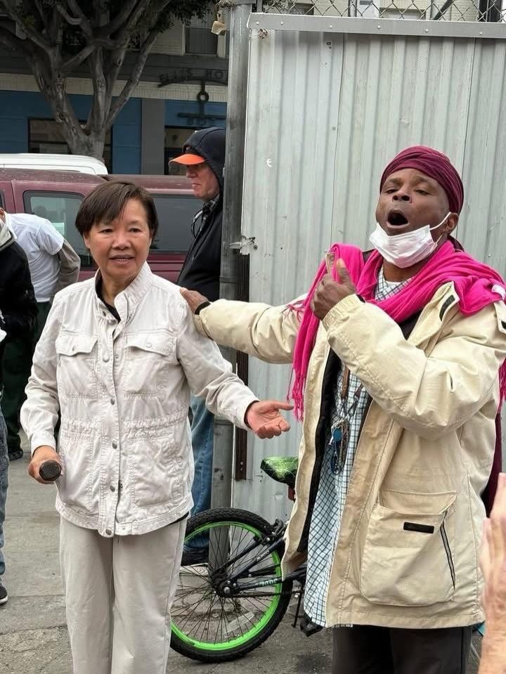A person with a pink scarf and face mask gestures while talking next to an individual in a beige jacket on a city street.