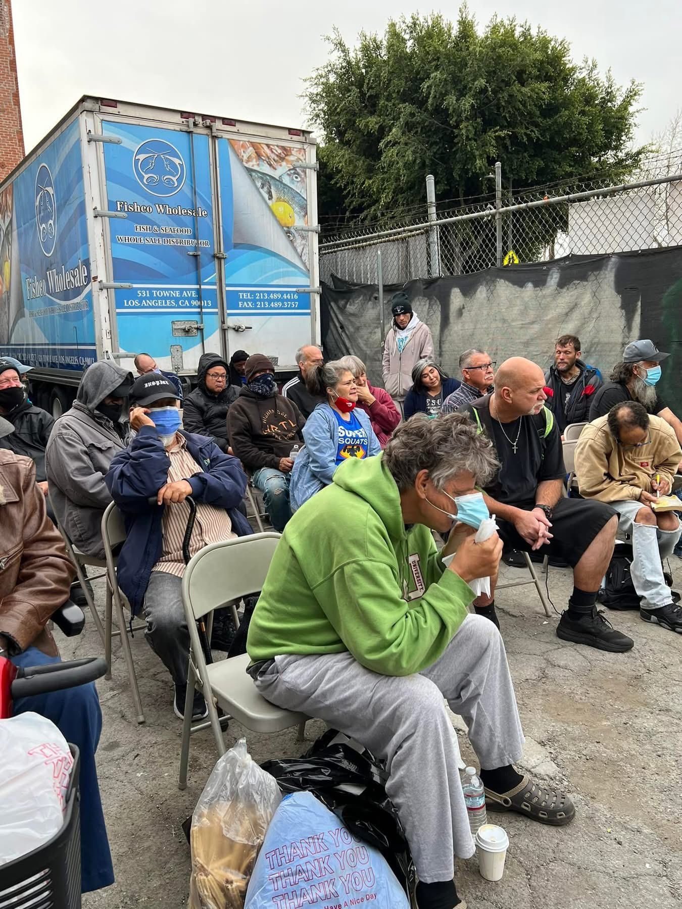 A group of people sit on folding chairs in an outdoor area near a blue semi-truck trailer, with some wearing face masks.