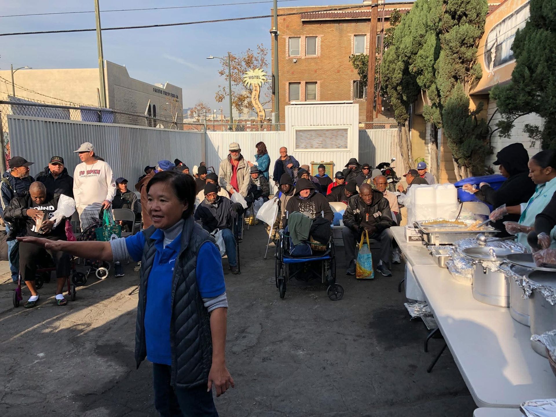 A volunteer points toward a line of people waiting for food in an outdoor urban setting with tables of supplies.