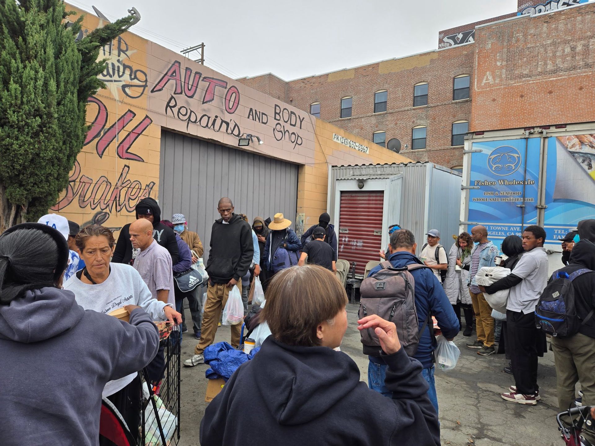 A crowd of people gathers for food distribution outside an urban auto repair shop.