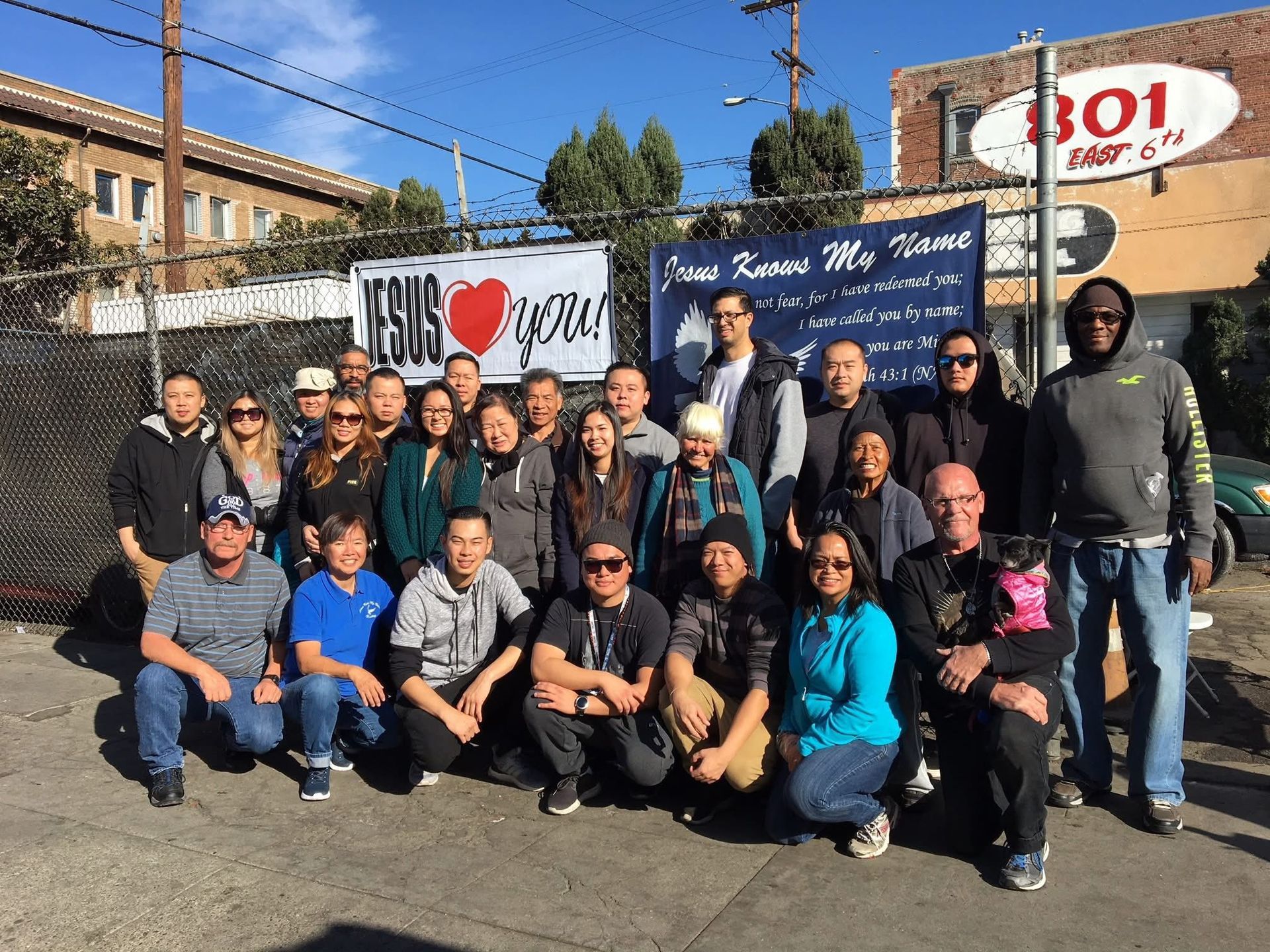 A large group poses in front of a chain-link fence decorated with banners promoting religious messages in an urban setting.