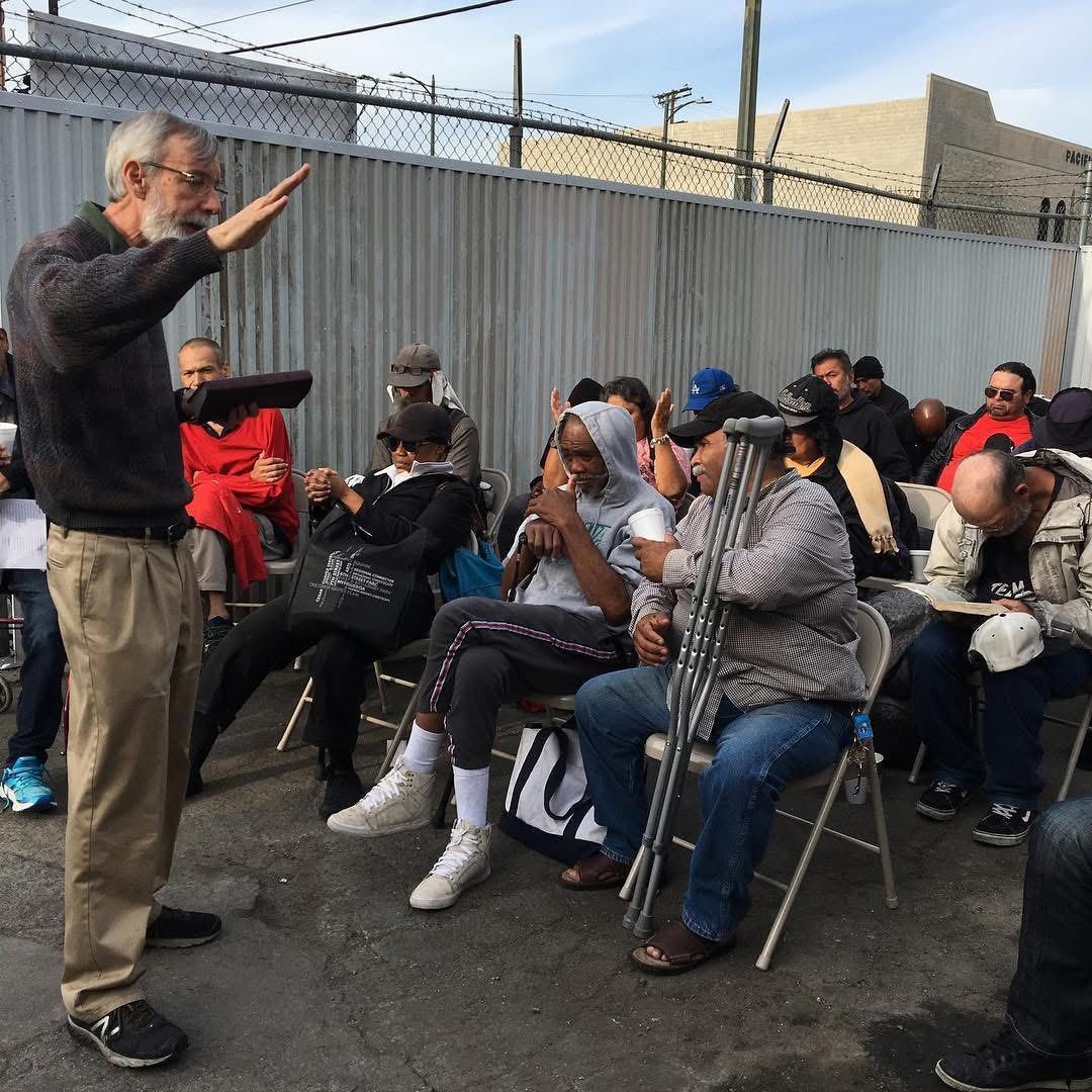 A man and a woman stand on a church stage, addressing an audience, with a live music band and cosmic background behind.