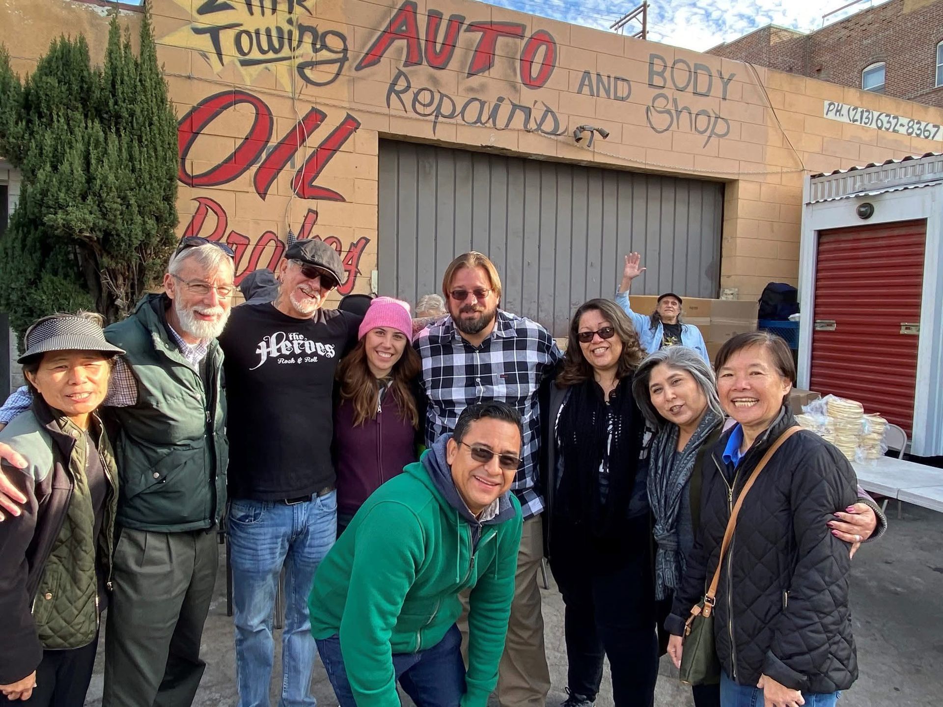 A group of nine people smiling together outside an auto repair shop with hand-painted signage on the brick wall.
