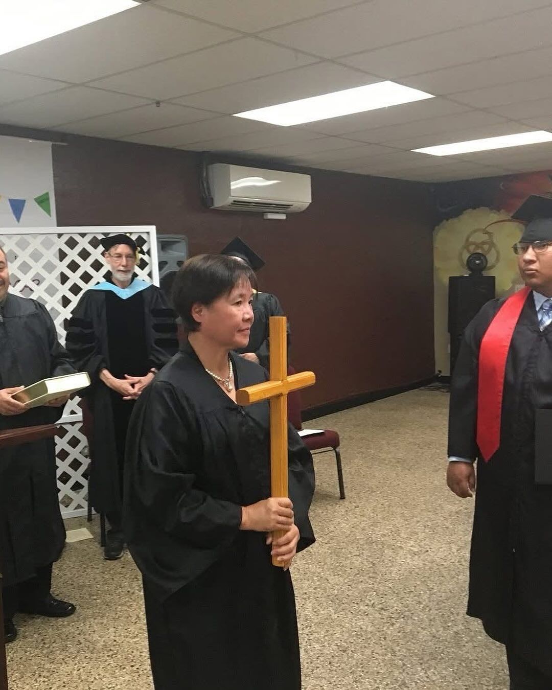 A person in graduation regalia holds a wooden cross during an indoor ceremony with attendees in the background.