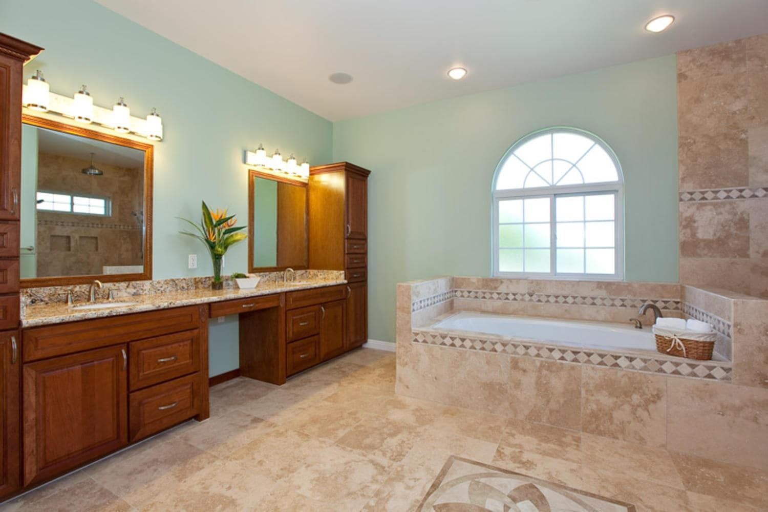 Spacious bathroom with light green walls, brown cabinetry, and beige tiled floor with a large arched window.