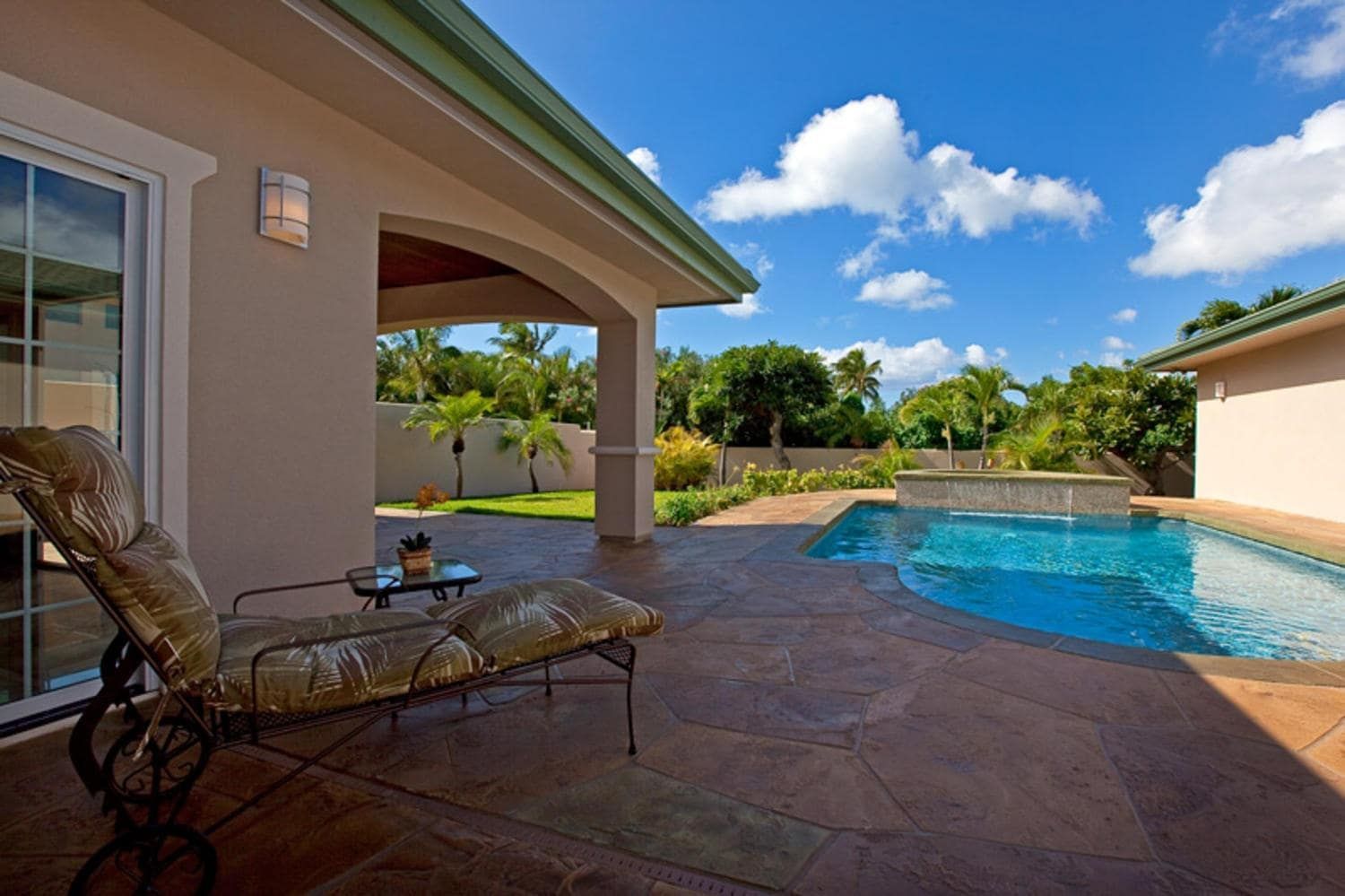 Lounge chair on a patio overlooking a pool and yard, under a blue sky with fluffy clouds.