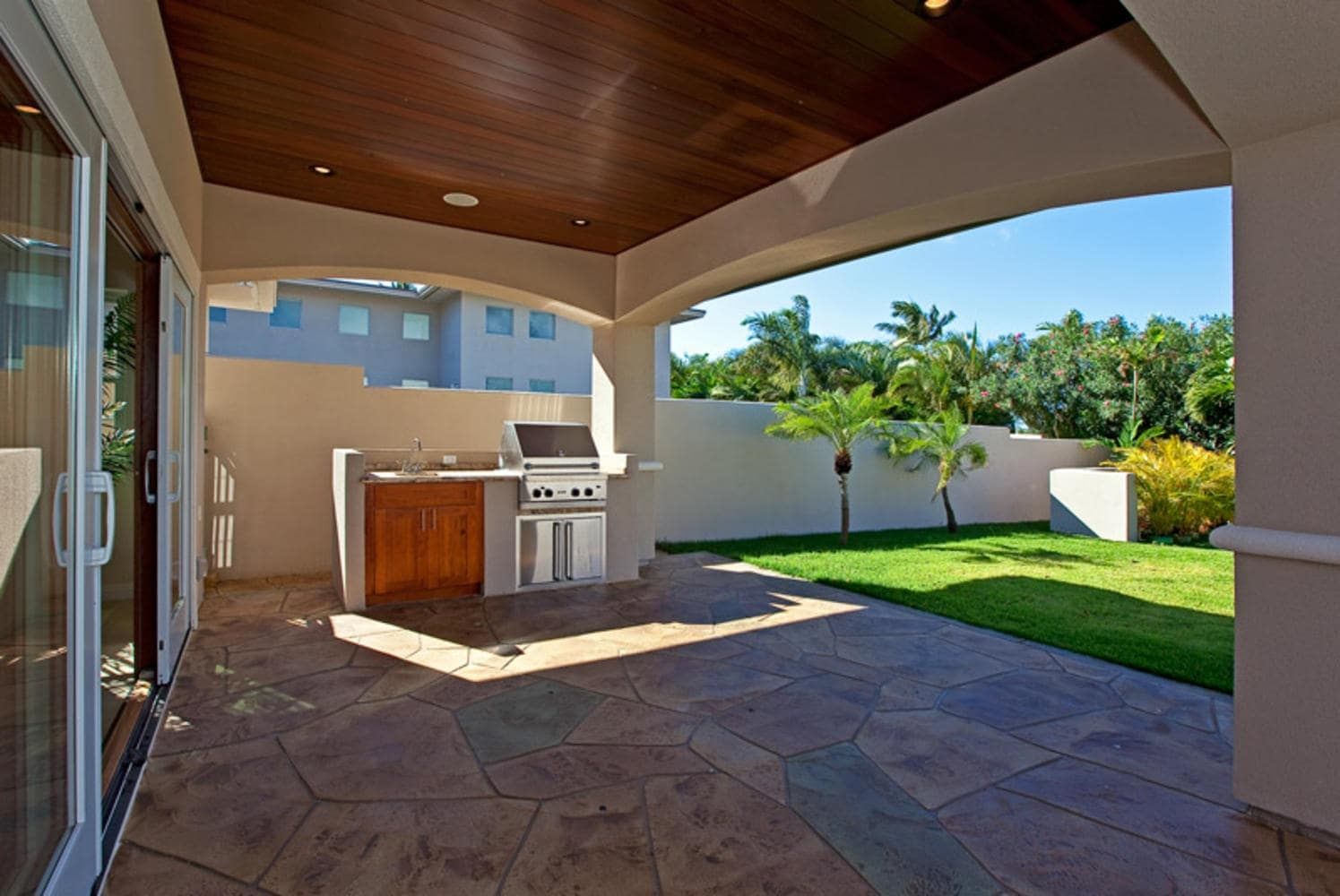 Patio with grill, wooden ceiling, stone floor, and view of yard with grass and trees.