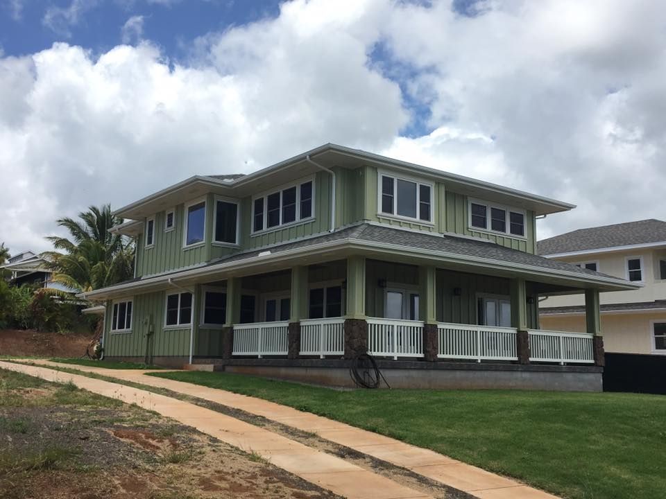 Green two-story house with a wrap-around porch, white railings, and a driveway on a grassy hill.