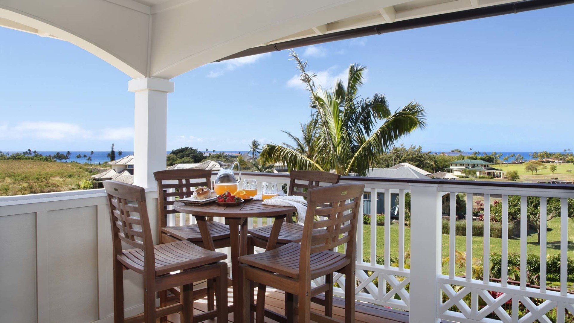 Balcony with wooden table and chairs, overlooking ocean and green landscape on a sunny day.