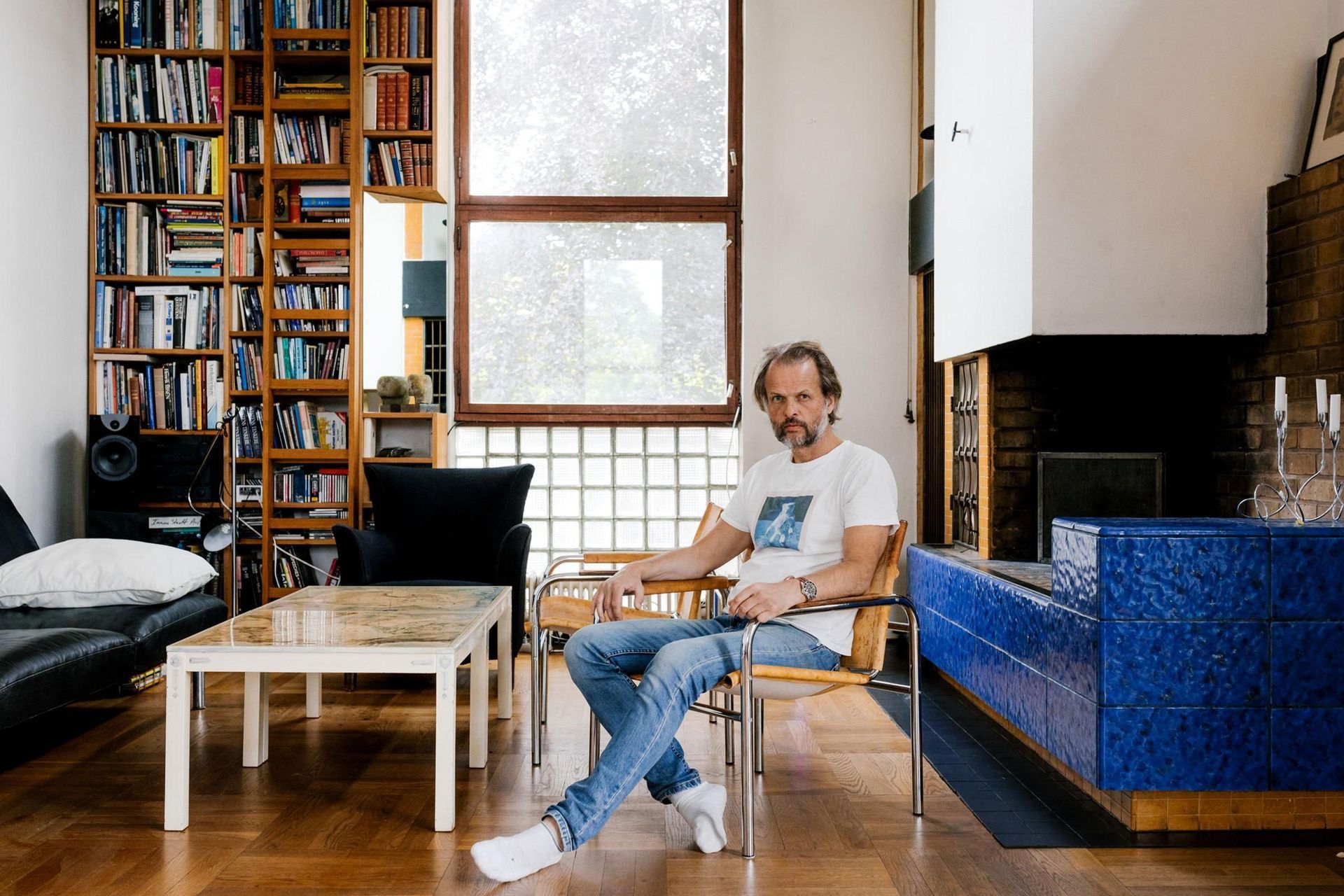 A man with a beard sits in a wooden chair within a bright living room, surrounded by a floor-to-ceiling bookshelf and a distinctive blue-tiled fireplace. Large windows provide natural light to the space, which features parquet flooring and modern furniture.