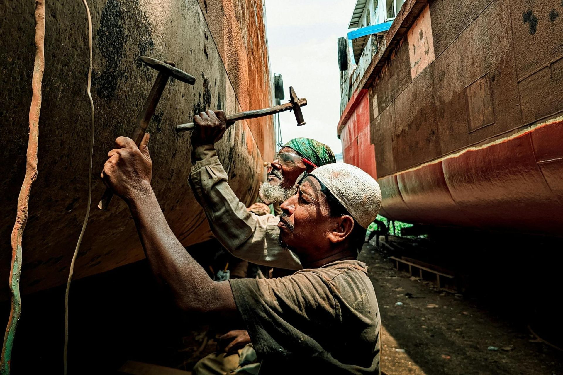 Two men are diligently working in a shipyard, using hammers to chip away at the weathered, rusty hul
