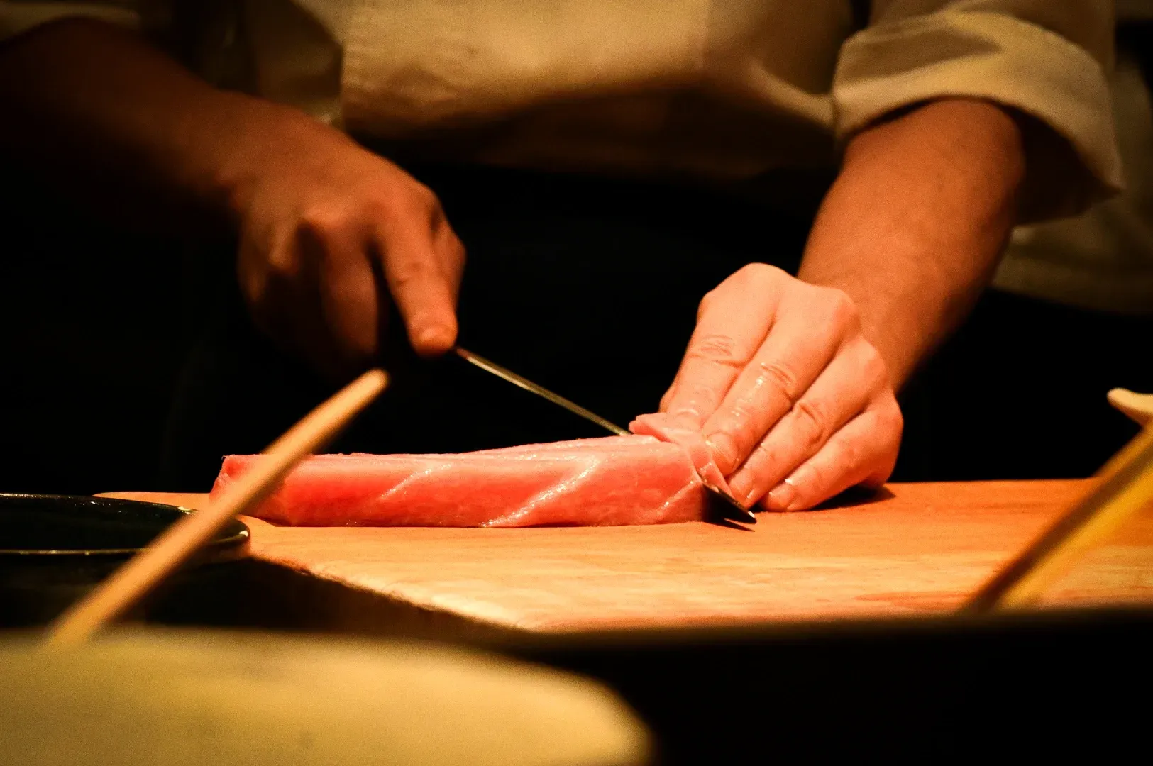 Tight close-up side-angle of chef slicing raw fish with precision knife skills on wooden board highl