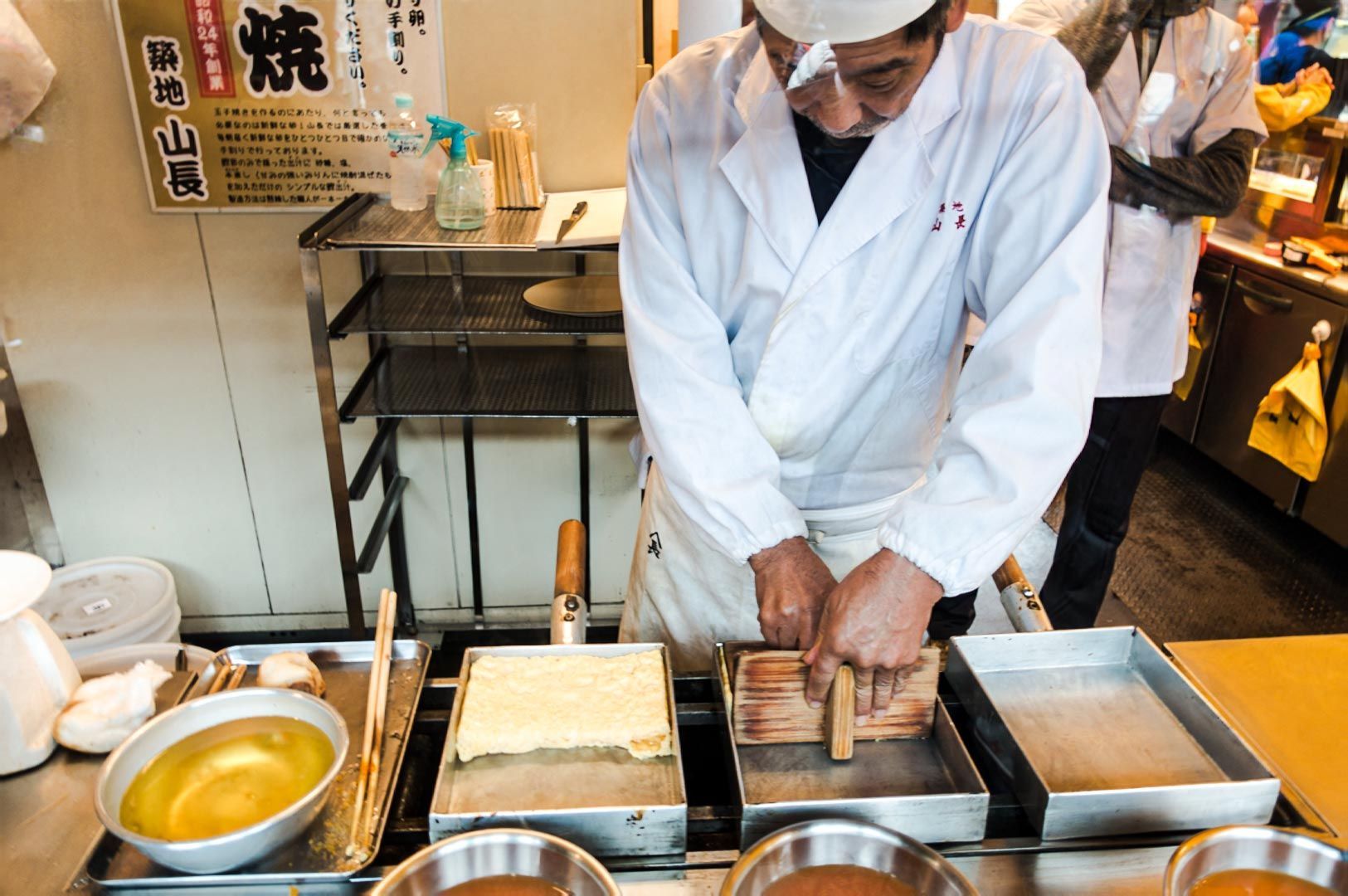 A chef preparing food at a market stall using wooden cooking tools, with trays and equipment arrange