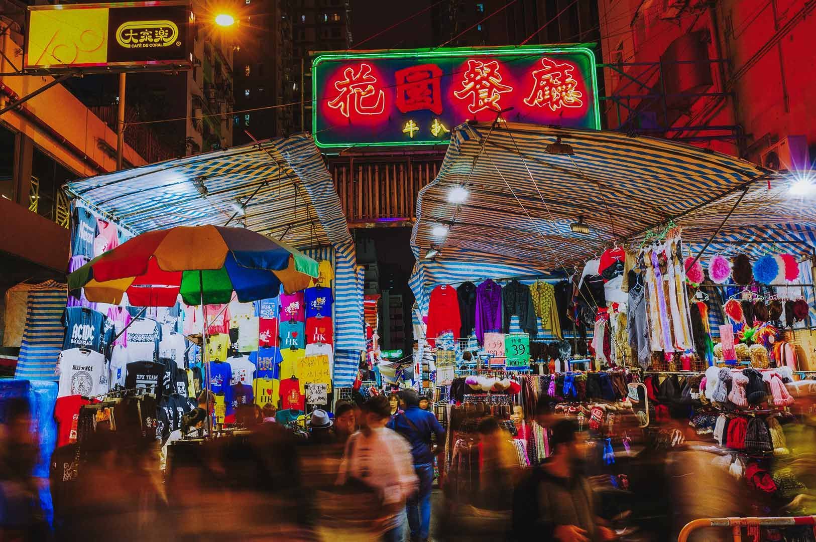 Busy night market with lit stalls selling clothes, striped tarpaulin covers, large umbrella, and neo