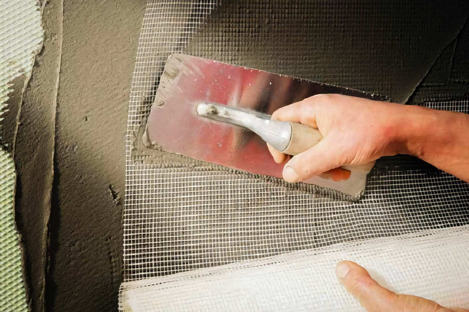 A close-up of a hand using a metal trowel to spread plaster or cement over a wall covered with reinf