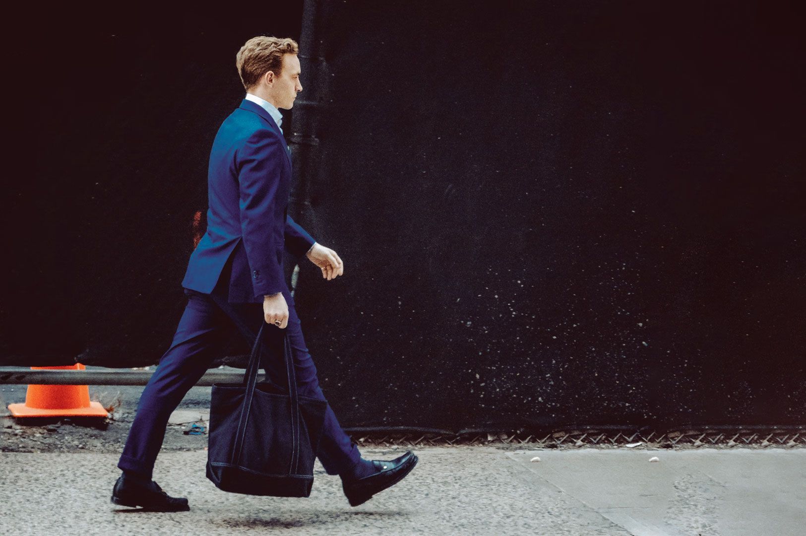 Person dressed in a formal suit walking briskly down a city street carrying a dark tote bag.