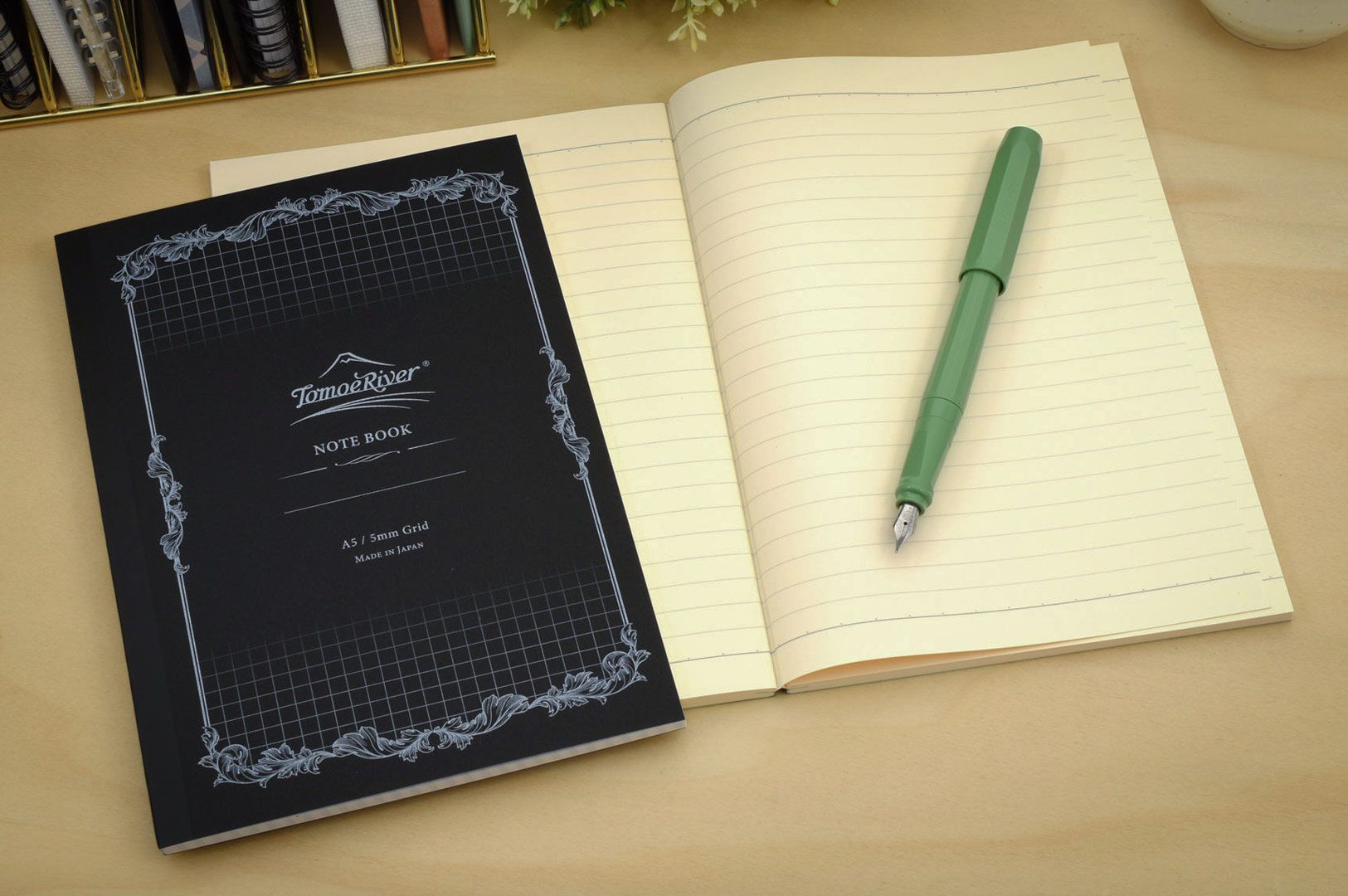 Open lined notebook with a green fountain pen beside a Tomoe River notebook on a wooden desk.
