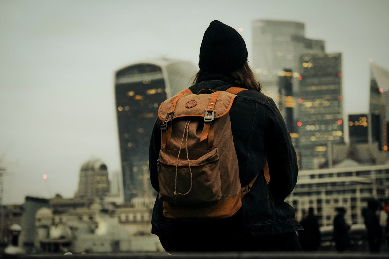 Traveler with a backpack looking at a modern city skyline with tall buildings on a cloudy day.