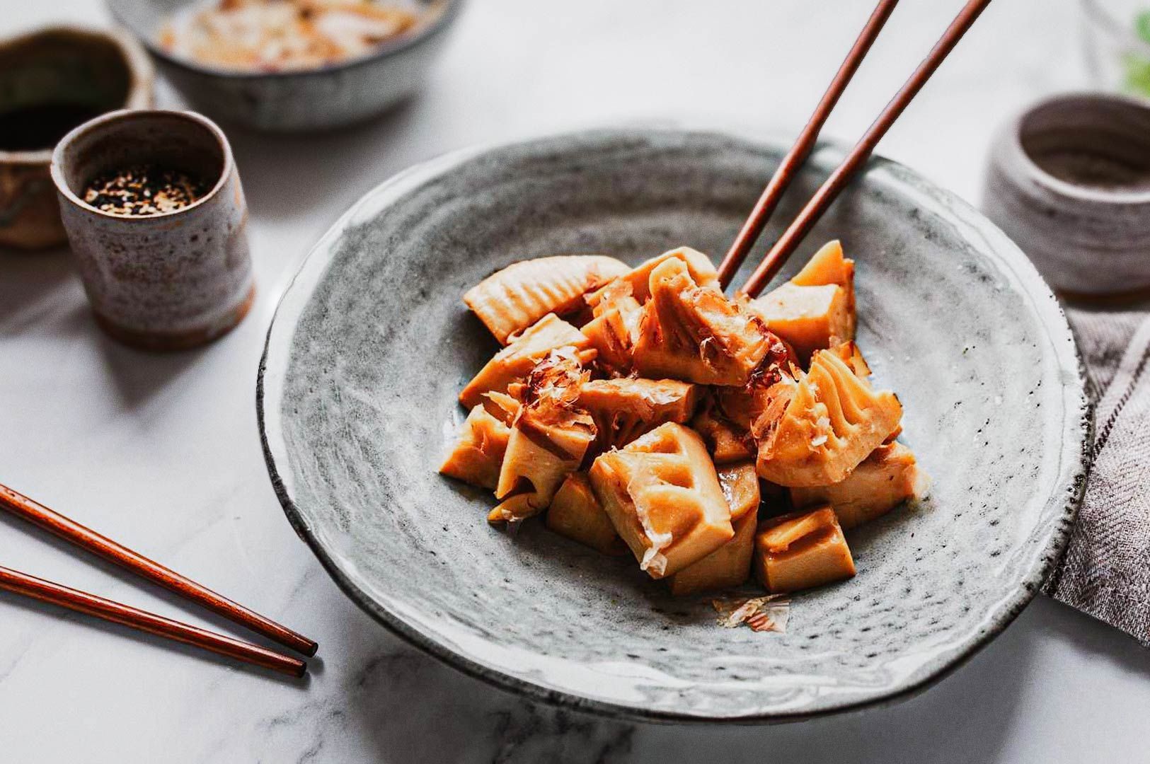 Close-up of a ceramic bowl filled with grilled bamboo shoots drizzled with sauce, with wooden chopst