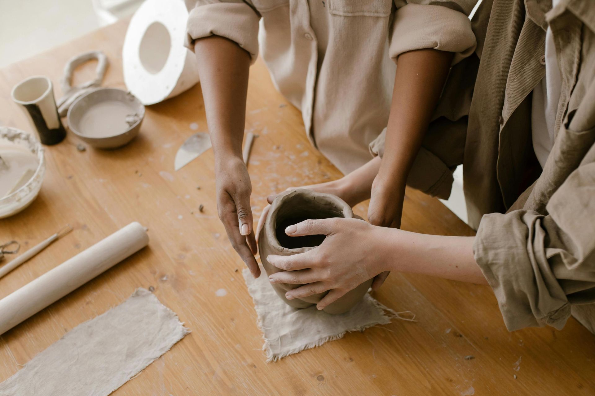 Two people are working together at a wooden table to hand-build a clay vessel, their hands covered in dust as they carefully shape the piece. The workspace is filled with various pottery tools, including a rolling pin, bowls of water, and scraps of fabric used for texturing.