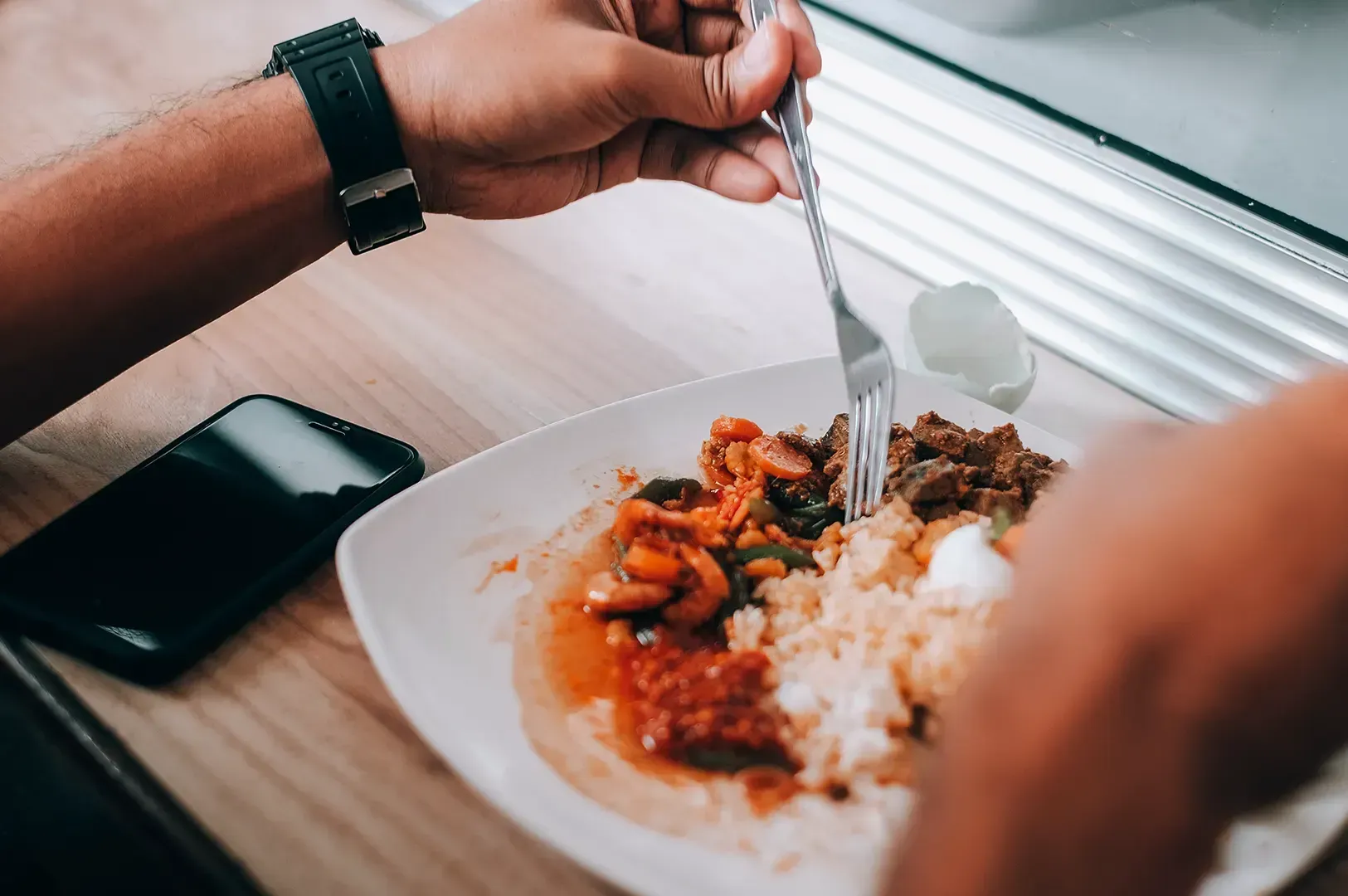 A close-up of a person eating a meal consisting of rice, meat, and vegetables at a table next to a smartphone, highlighting the personal, individual dining choices.