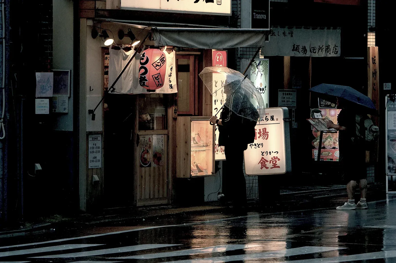 A rainy evening scene in a Japanese alleyway featuring a person with a transparent umbrella standing outside a traditional restaurant, capturing the beauty of slow, intentional urban exploration.