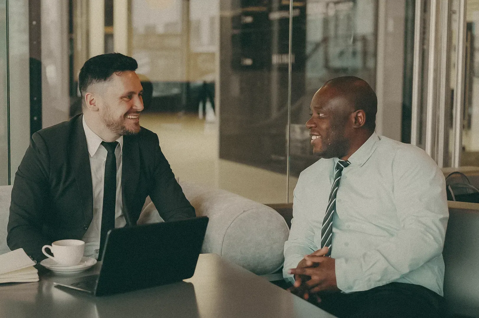 Two professionals having a  in a modern office lounge, seated with a laptop and coffee on the table.